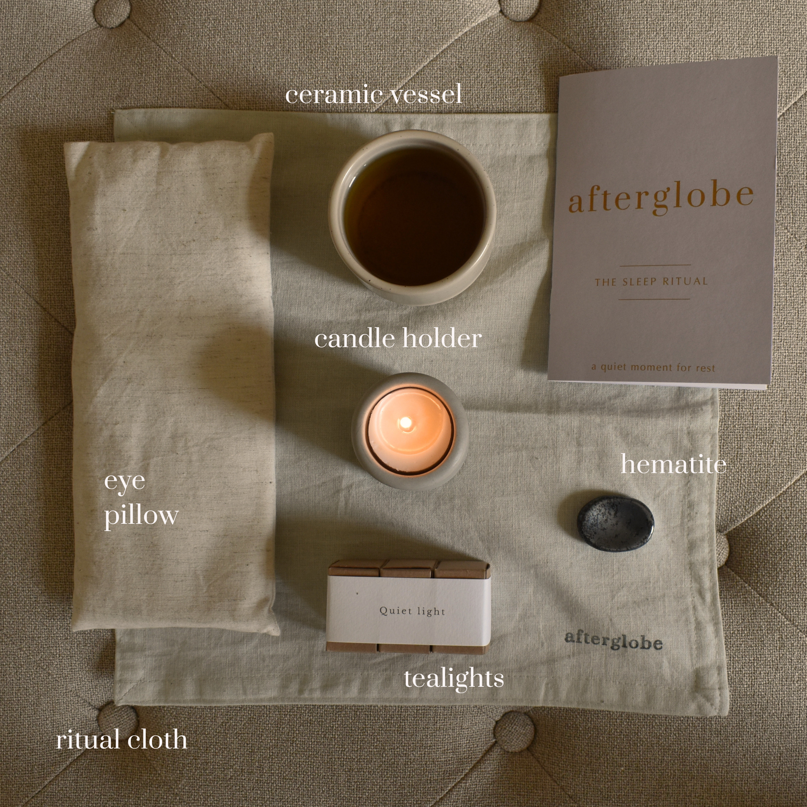 Collection of items including a ceramic vessel, candle holder, eye pillow, ritual cloth, and a book titled 'afterglobe' on a textured surface.